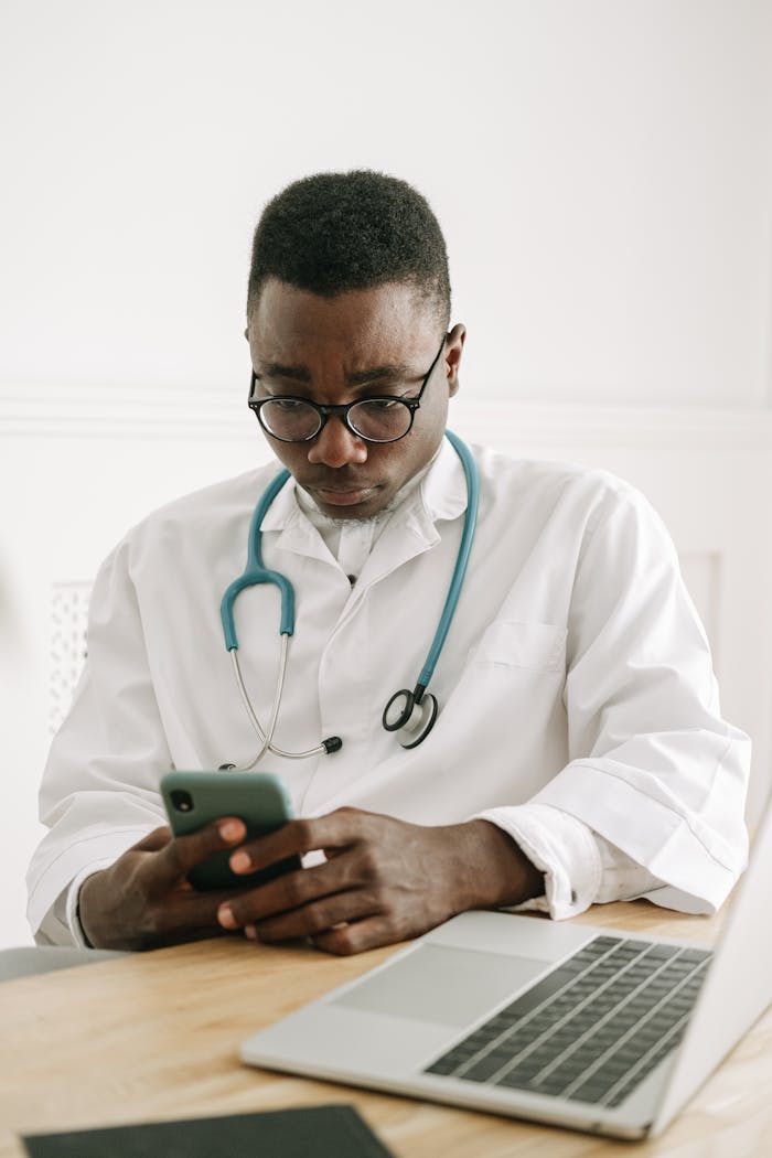A professional doctor in a medical gown using a smartphone at a workplace desk with a laptop and stethoscope.