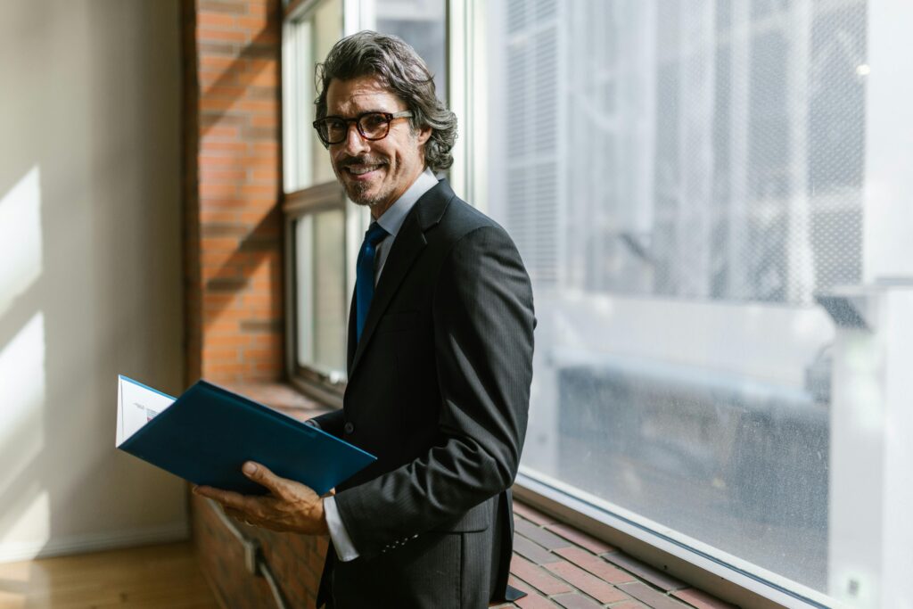 Smiling businessman in a black suit holding a folder by a window indoors.